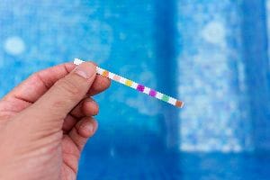 Hand Holding A Pool Chemical Testing Strip Near A Pool.
